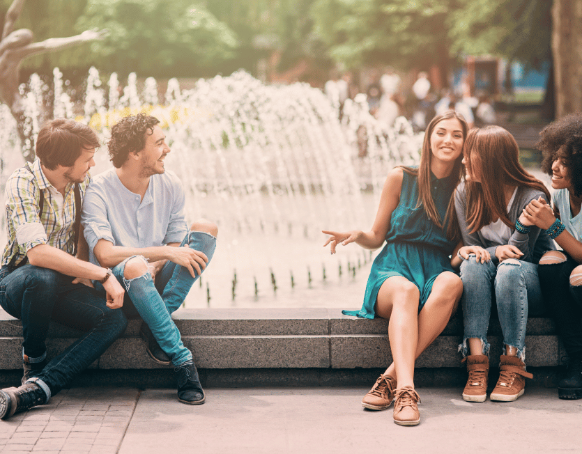 Zwei Männer beim Frauen ansprechen – lockere Stimmung auf einem Springbrunnen im Park mit drei lachenden Frauen.