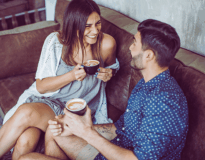 Ein Mann flirtet in einem Café in Ulm mit einer attraktiven Frau. Beide sitzen entspannt auf einer Couch und lachen miteinander beim Date.