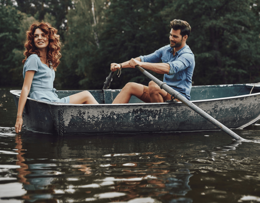 Paar sitzt in einem Ruderboot auf einem ruhigen See; der Mann rudert, die Frau sitzt entspannt am Bootsrand und berührt das Wasser.